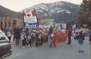 Groupe de jeunes enfants en tenue de ski défilant dans une parade en tenant un grand drapeau portant l’inscription « Nancy Greene Ski League ».