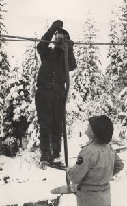 Man standing on a ski chair attached to a thick wire rope while another man holds the chair at the bottom. The man standing on the chair is using a tool to secure the chair to the rope.