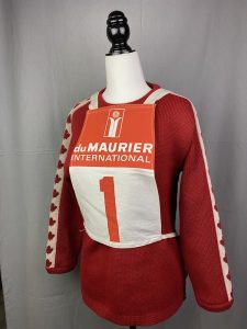 Red sweater with maple leaves on the sleeves and an orange and white racing bib over the body.