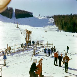 Skiers in line for the Poma Lift and skiing down a run on right of the lift.