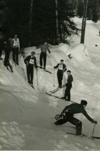 Six male skiers watching a seventh skier come down a ski run.