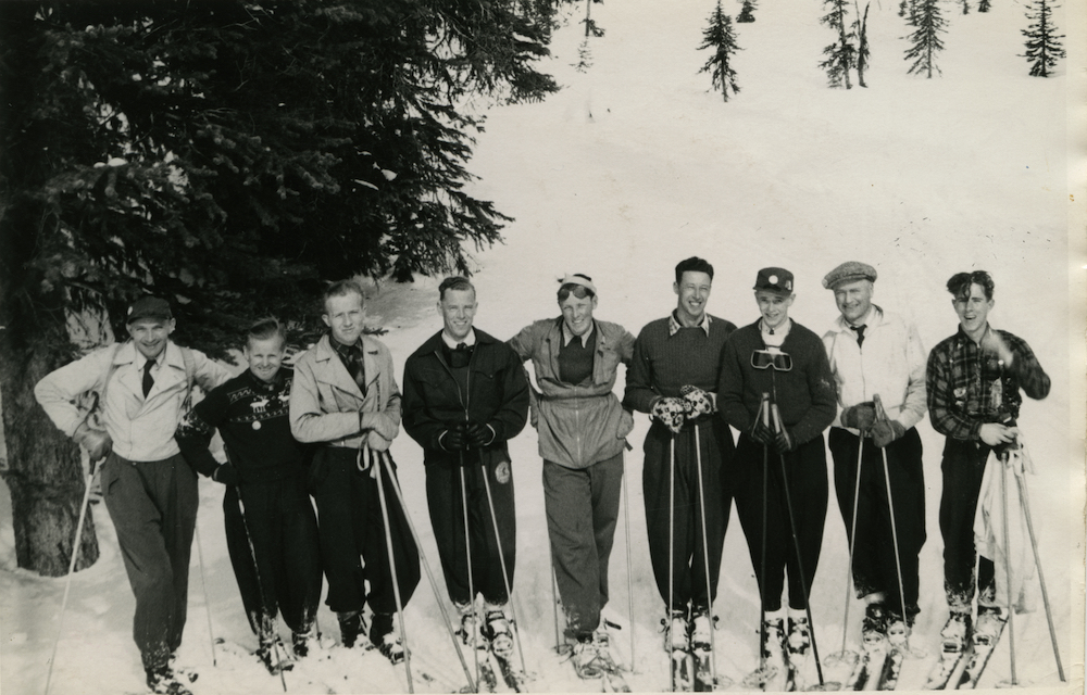 Group of nine men on skis photographed on a snow-covered mountain slope with trees in the background.
