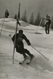 Black and white photo of two skiers watching a young male skier going through a slalom gate.
