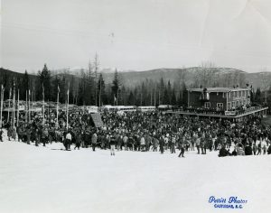 Black and white photograph of a large crowd of spectators standing on snow looking at a podium.