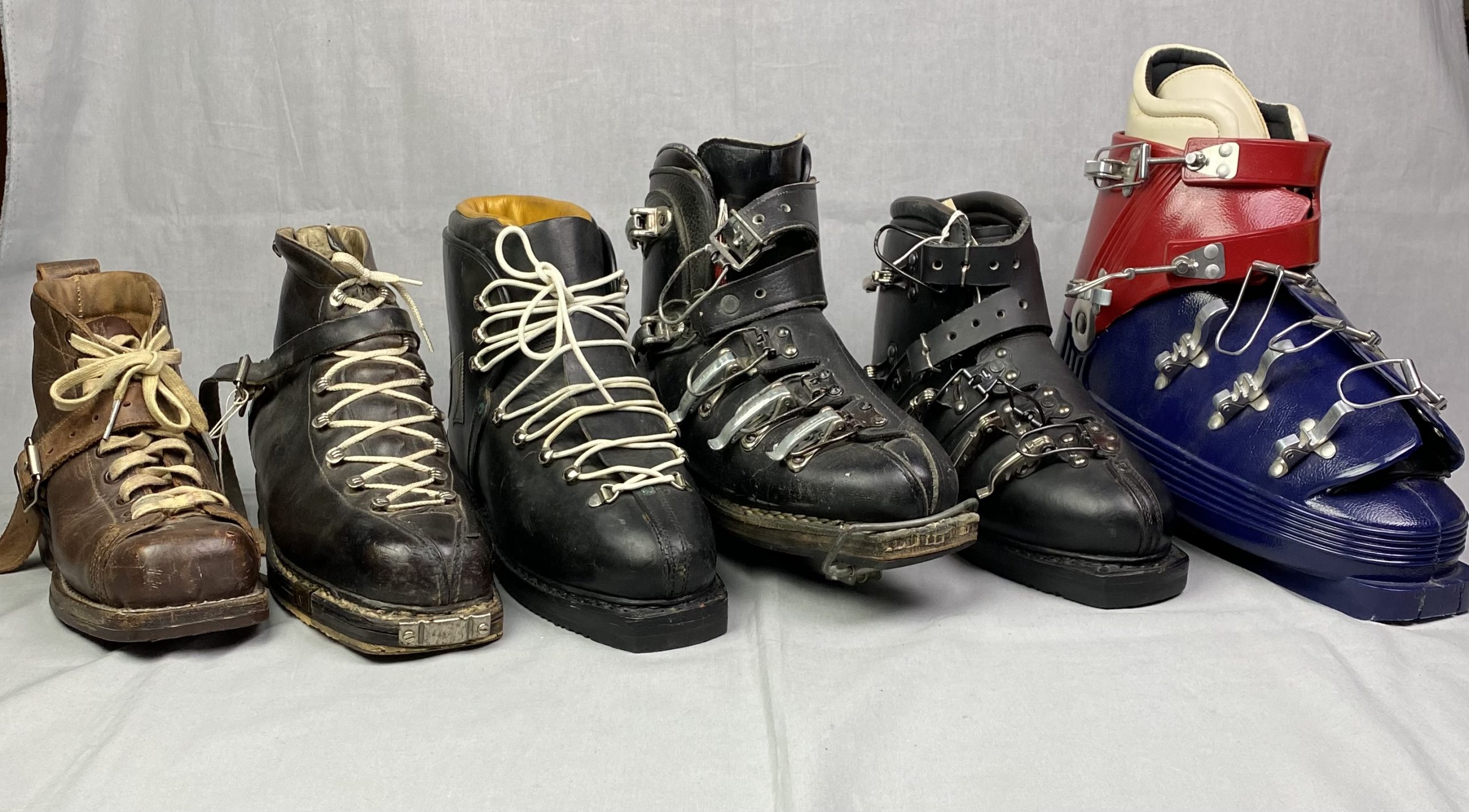 Six ski boots in a diagonal row showing a progression of styles through the decades. The three pairs are made of leather and have laces, the next two pairs are made of leather and have buckles, and the last pair is made of plastic and has buckles.