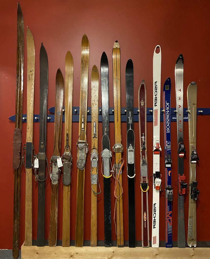 Fifteen different sized skis leaning against a red wall, showing a progression of equipment over the decades, from wooden skis to plastic skis to fibreglass skis.