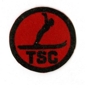 Round red felt patch with a black border and a graphic of a ski jumper over the letters TSC in the centre.