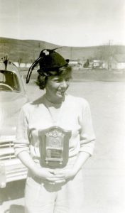 Young woman wearing a plaque as a medal around her neck with her hands crossed underneath standing in front of a car.