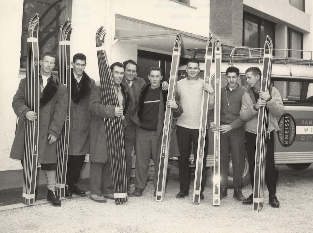 Eight men posing for a photo while holding up stacks of skis.
