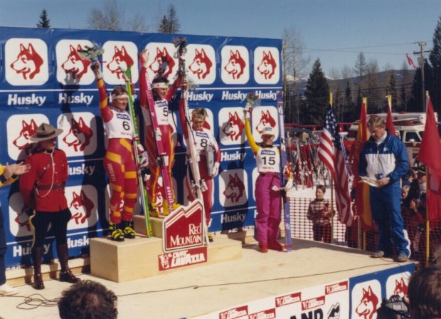 Four female skiers on a podium holding up their flowers in front of a Husky banner.