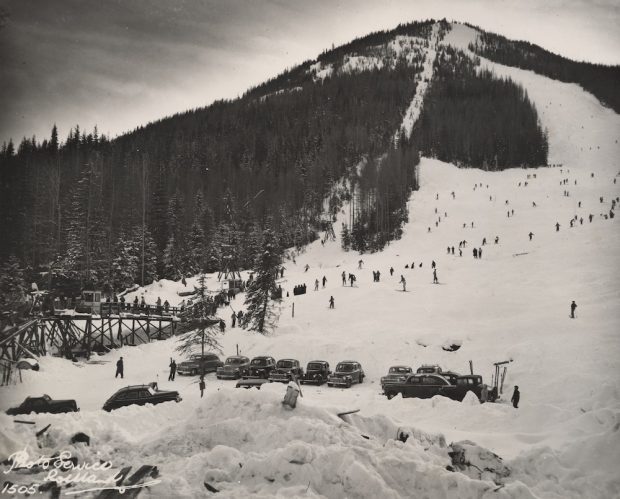 A black and white photo showing the chairlift, ski slope, and parking lot of Red Mountain. Many skiers are skiing and standing in line for the lift and there are fourteen cars visible in the parking lot.