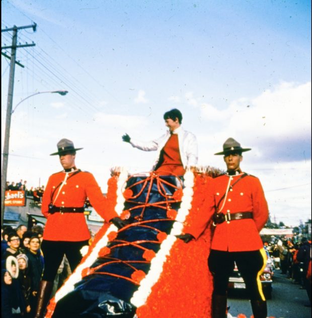 Nancy Greene sits atop a large red ski boot parade float waving at the crowd with two Mounties walking alongside.