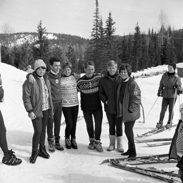 Black and white photo of a group of 6 people including three men and three women standing on the ski hill surrounded by ski gear.