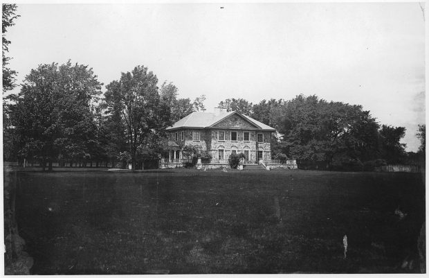 Black and white photograph of Grantham Hall, a large two-story stone house surrounded by a stone porch, trees, and shrubs.