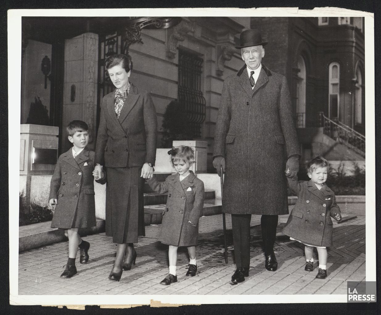 Black and white photograph of two parents and their three young children walking hand in hand on a sidewalk, in front of stone buildings.