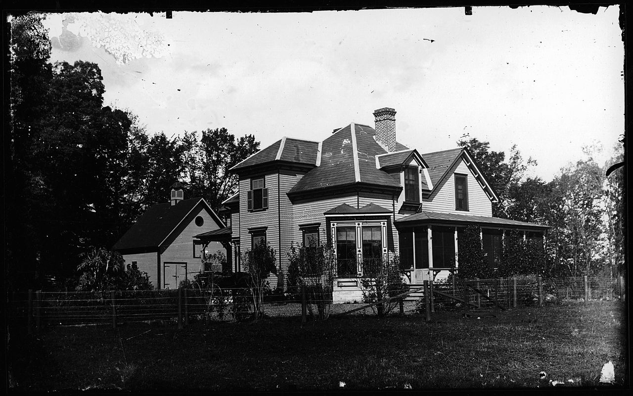 Black and white photograph of a large two-storey wooden house, decorated along the windows and surrounded by a fence. A barn is visible behind the house.