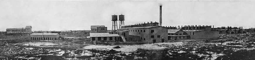 Photographie panoramique en noir et blanc d’une usine comprenant divers bâtiments, deux réservoirs et une cheminée.