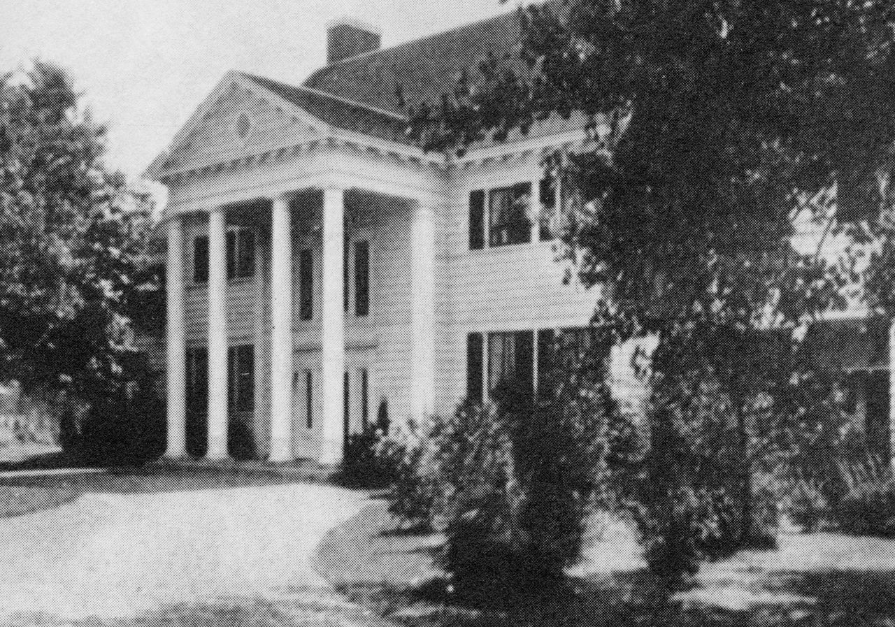 Photographie en noir et blanc d’une maison de style colonial en bois de deux étages avec des volets aux fenêtres et des colonnes entourant l’entrée de la demeure.