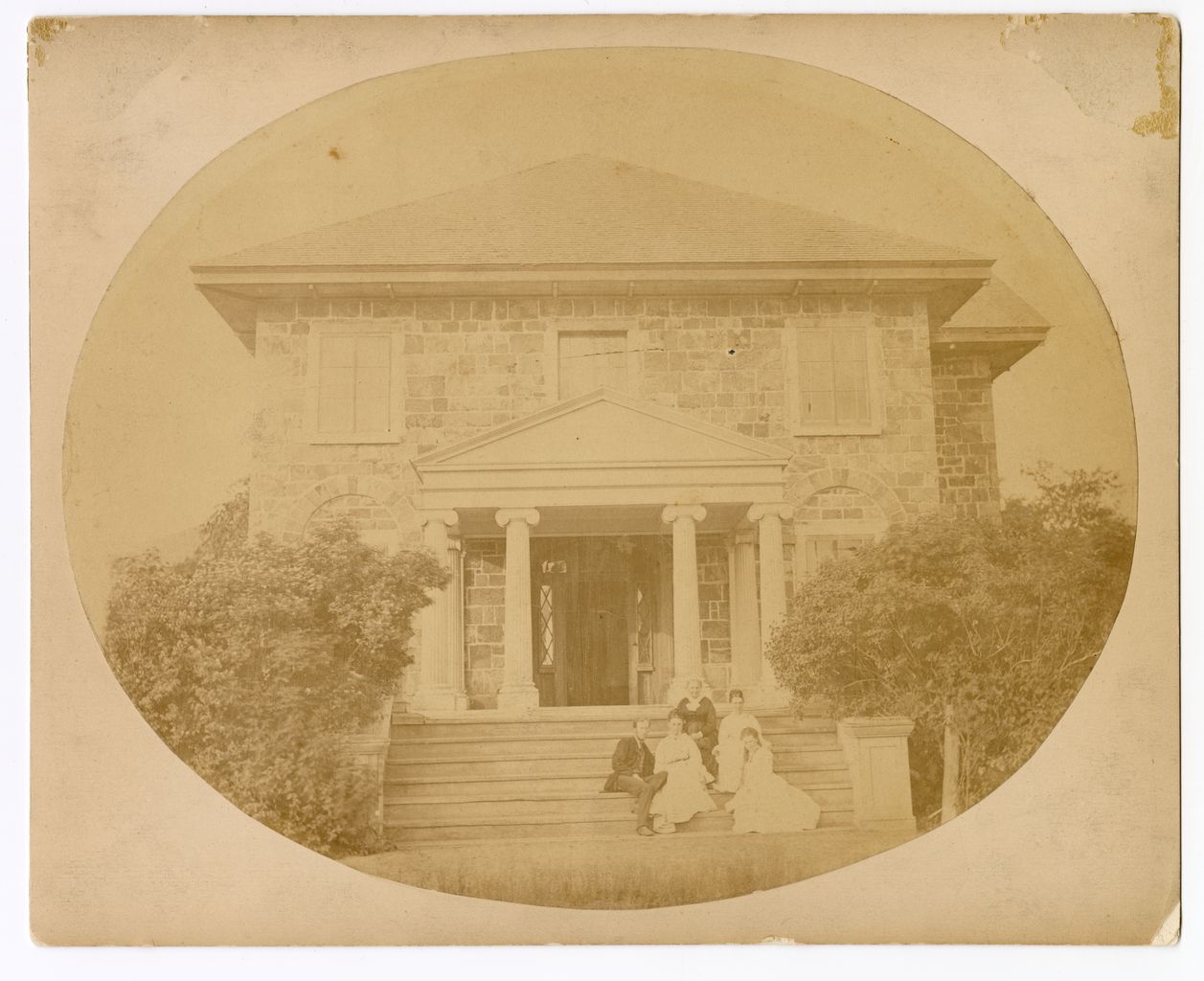 Black and white photograph of the facade of Grantham Hall, a large two-story stone house with a porch supported by columns and family members posing on the steps.