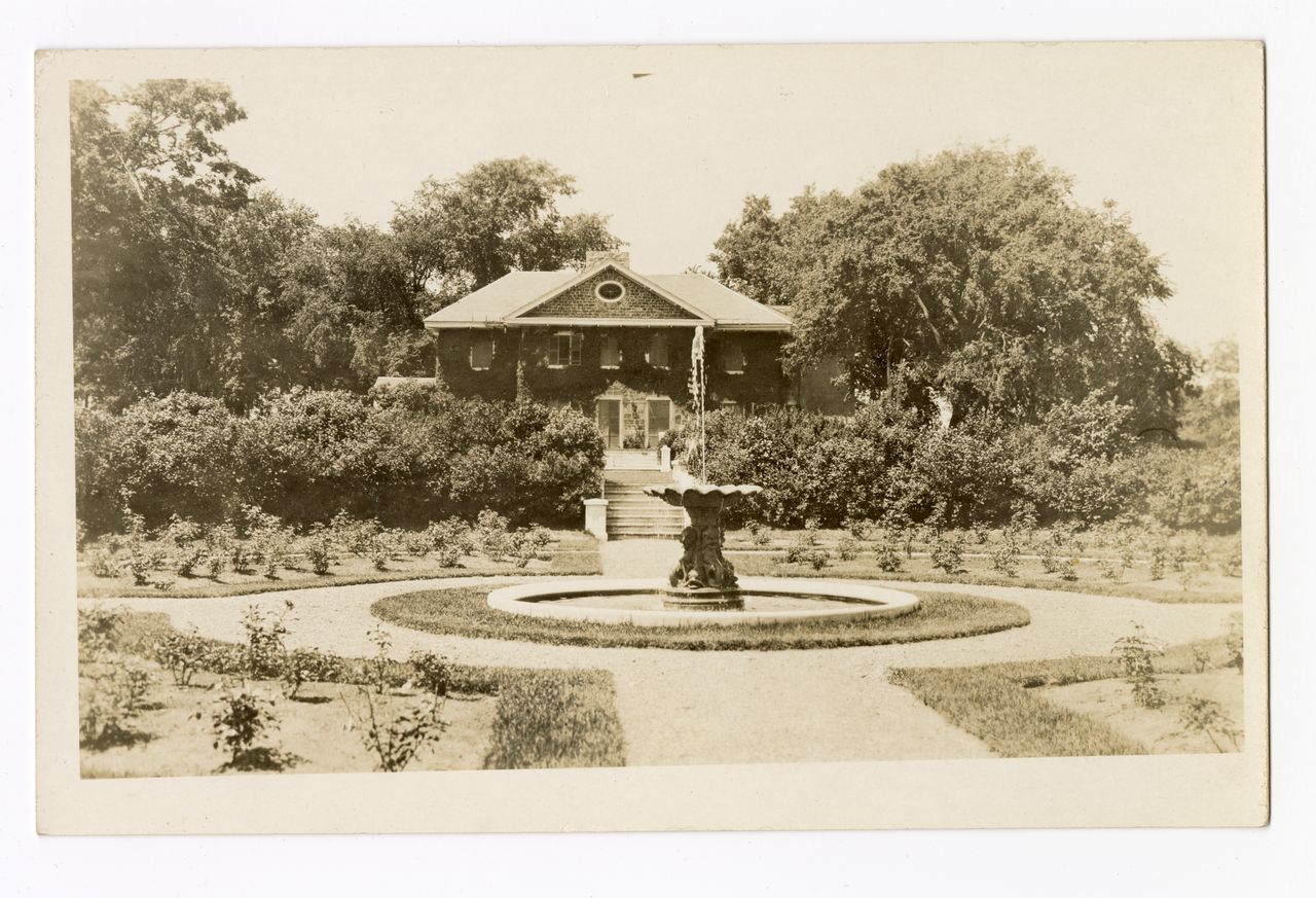 Black and white photograph of a circular garden of shrubs surrounding a fountain in front of a large two-story stone house covered in climbing plants and with a staircase leading to the garden.