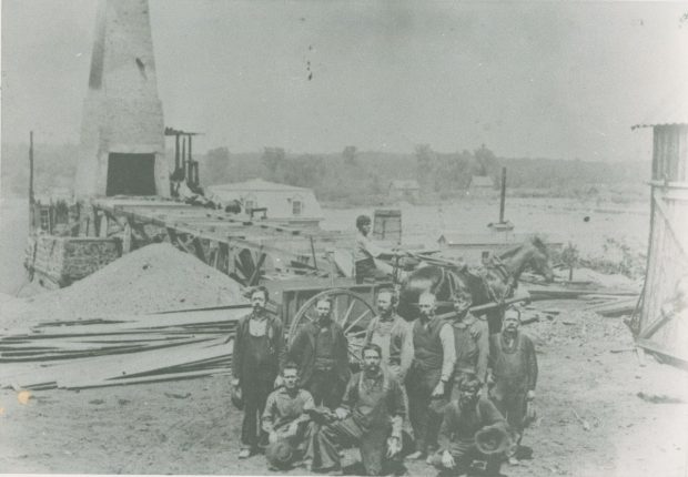 Black and white photograph of several men in front of a stone and brick blast furnace at the McDougall ironworks on the shore of the Saint Francis River.