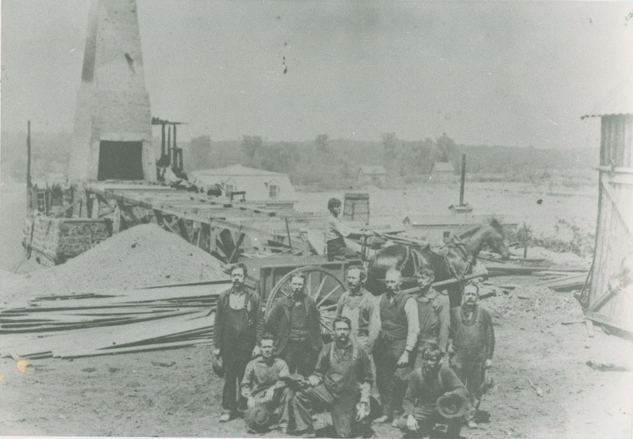 Photographie en noir et blanc de plusieurs hommes devant un haut fourneau en pierre et en brique des forges McDougall sur la rive de la rivière Saint-François.