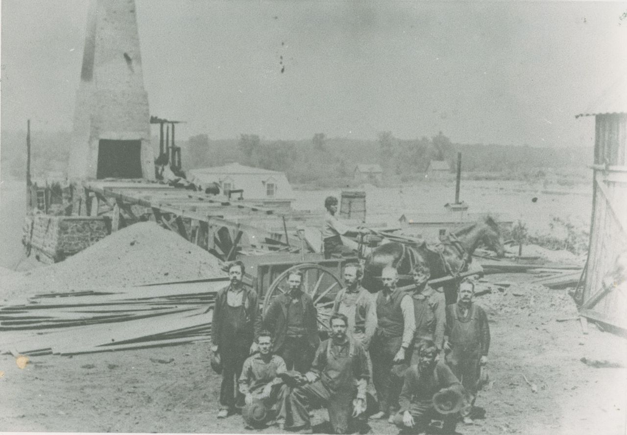 Black and white photograph of several men in front of a stone and brick blast furnace at the McDougall ironworks on the shore of the Saint Francis River.