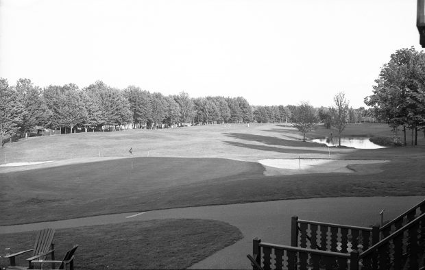 Black and white photograph showing a view of the porch of the golf course clubhouse. Golf flags and a resting area for golfers is visible.