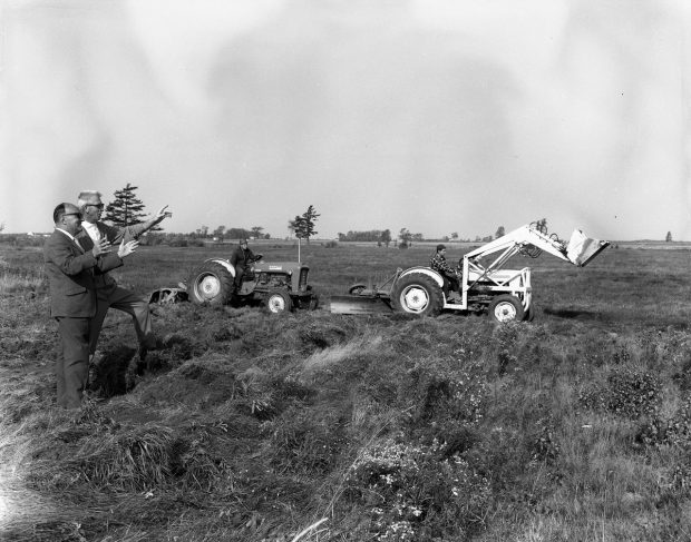 Black and white photograph of two men overseeing the start of construction of the golf course on vacant land. Behind them, two men operate machinery.
