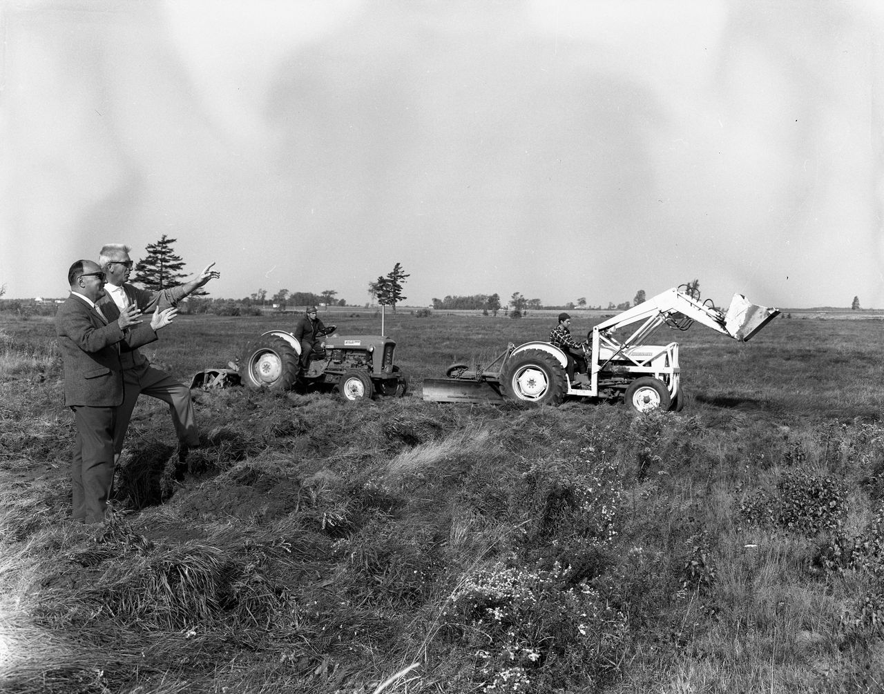 Photographie en noir et blanc de deux hommes supervisant le début des travaux du terrain de golf sur un terrain vague. Derrière eux, deux hommes manœuvrent de la machinerie.