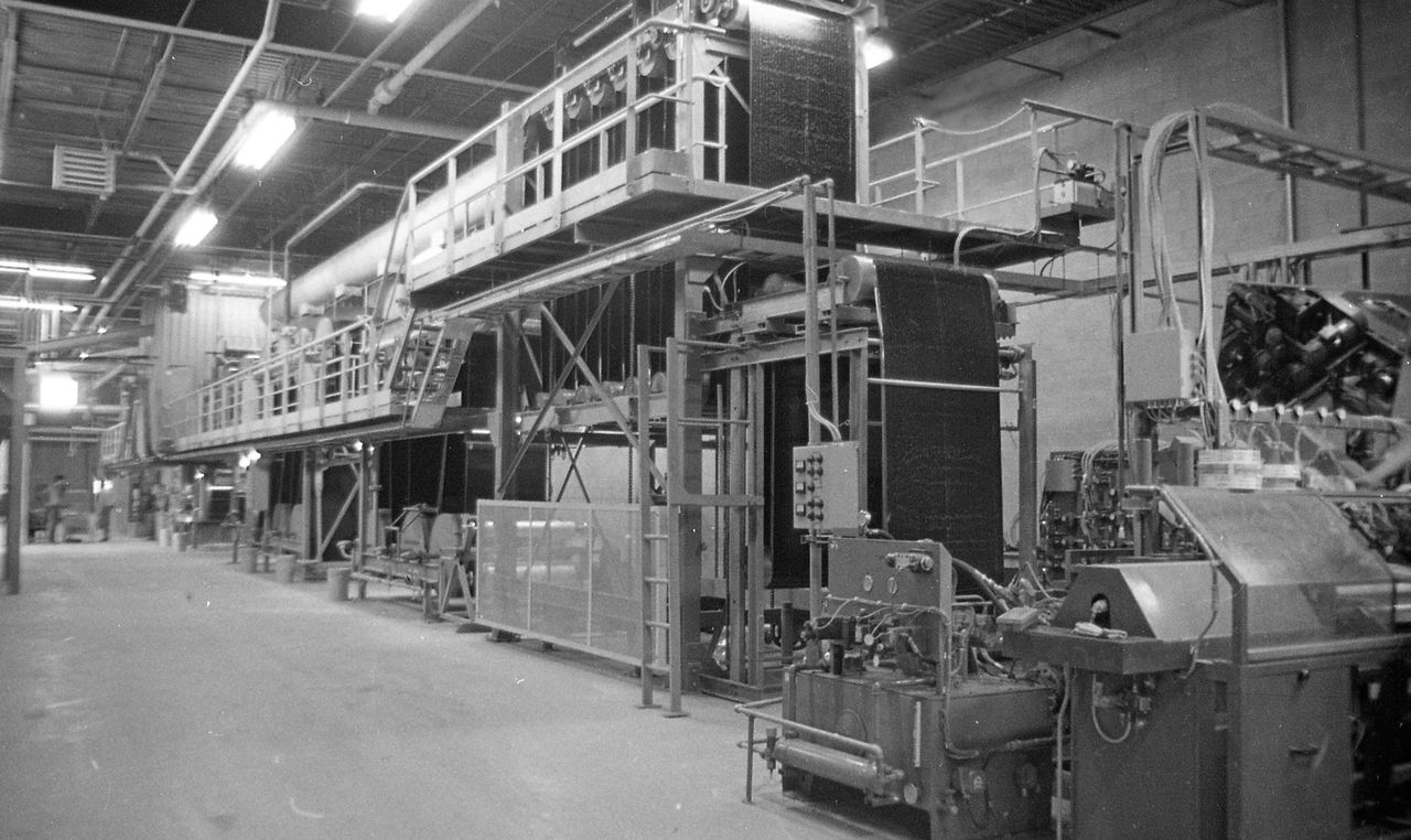 Black and white photograph of the interior of the Soprema factory. It shows several machines in a concrete room.