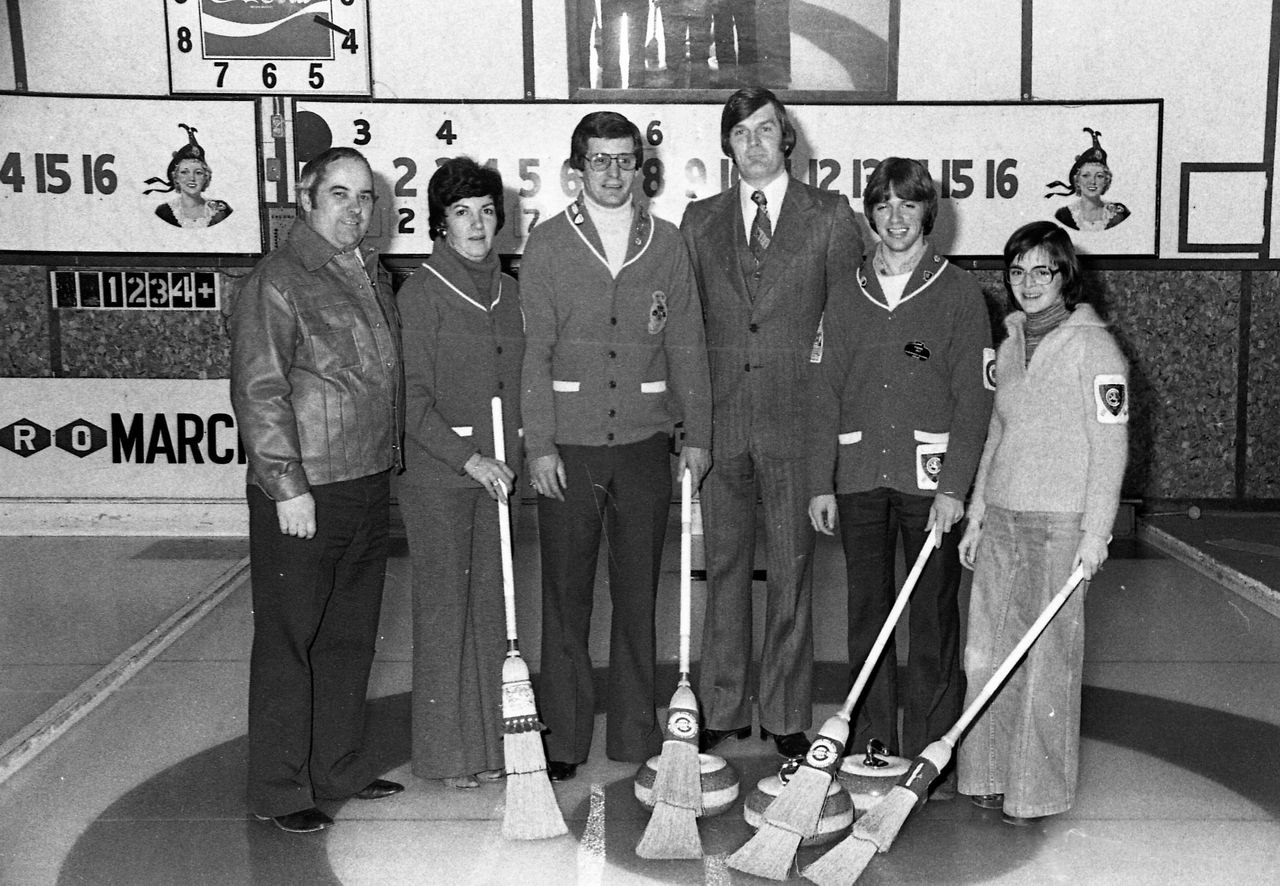 Photographie en noir et blanc d’un groupe de personnes de tous âges debout sur une piste de curling, des balais à la main.