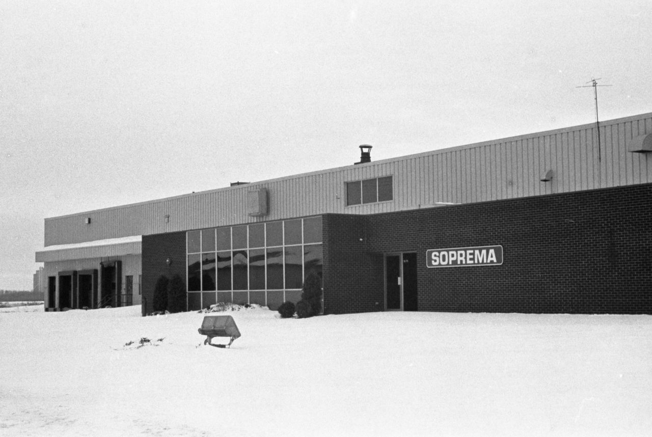 Black and white photograph of the facade of the Soprema factory. It shows the sign on the large glass bay window and three loading docks.