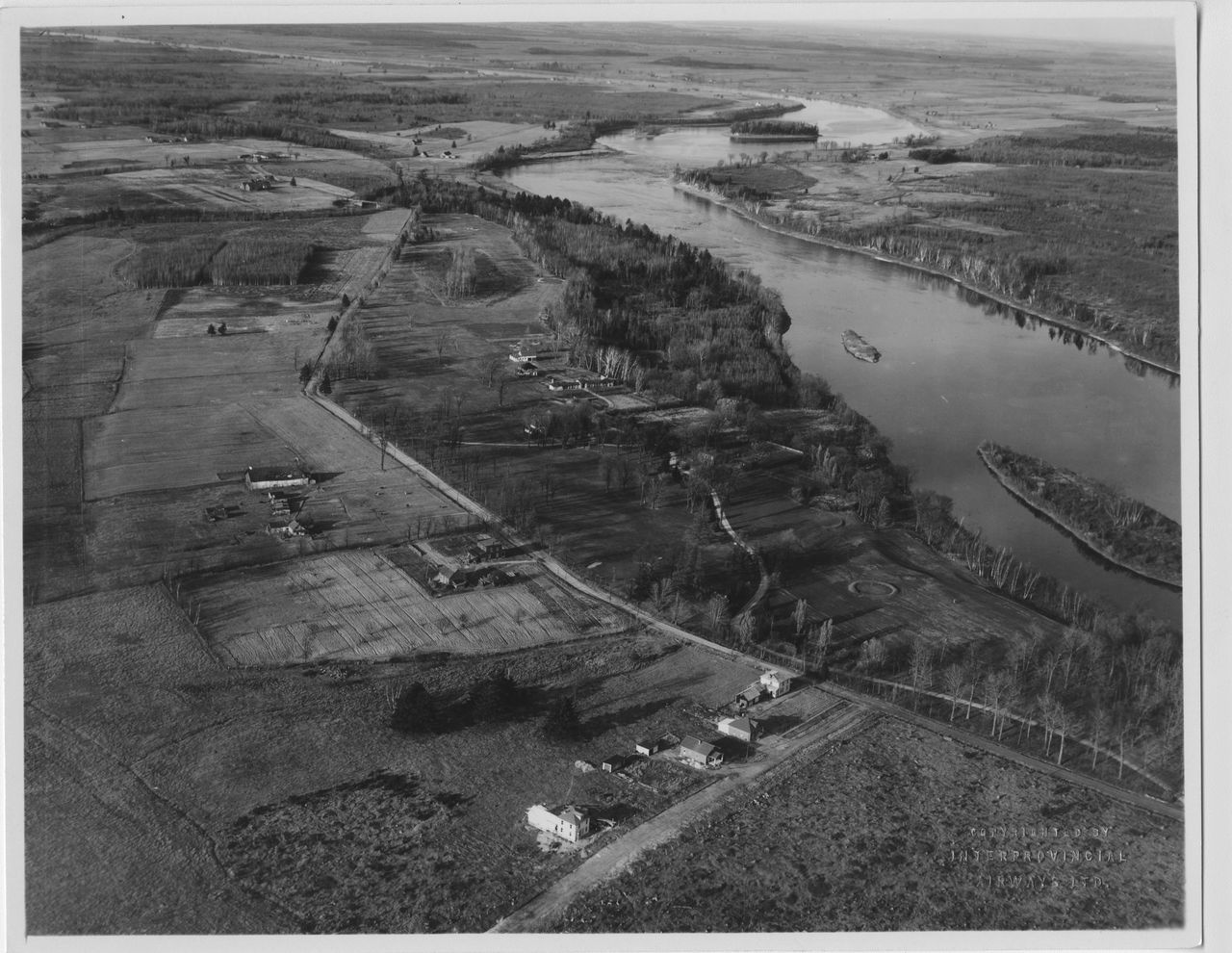 Photographie en noir et blanc démontrant une vue aérienne du parcours de golf aménagé le long de la rivière Saint-François. On y voit également plusieurs maisons dispersées dans les champs ceinturant le terrain de golf.