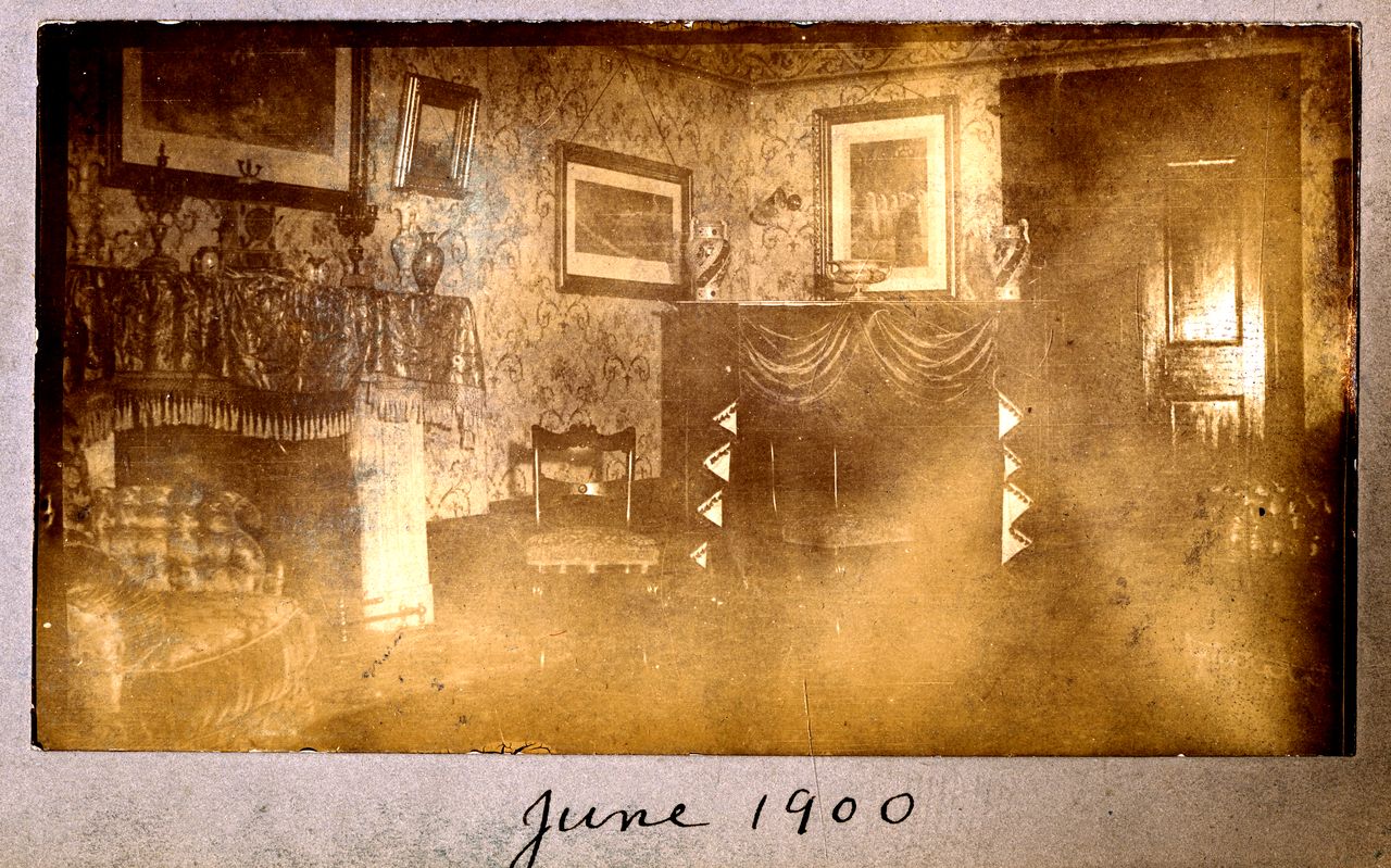 Photographie en noir et blanc du salon du Grantham Hall décoré par des cadres sur les murs et des draperies sur les meubles. On y voit une cheminée, des chaises et un fauteuil.