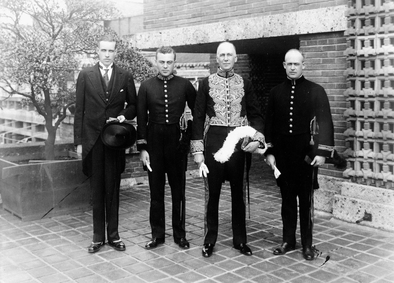 Photographie en noir et blanc de plusieurs hommes en uniforme et en complet devant un arbre en pleine ville.