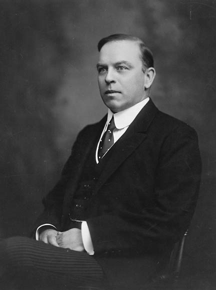 Photographie en noir et blanc d’un homme posant assis, vêtu d’un costume-cravate.