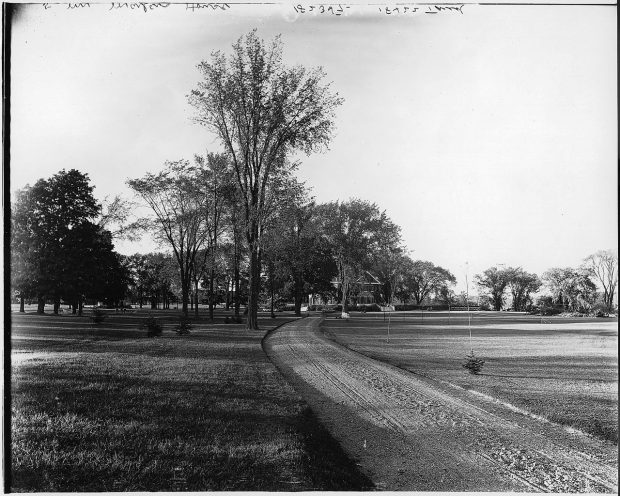 Black and white photograph of a dirt road lined with trees leading to a large two-story stone house.