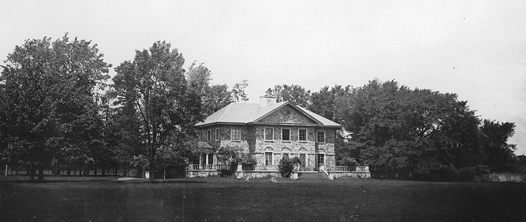 Black and white photograph of Grantham Hall, a large two-story stone house surrounded by a stone porch, trees, and shrubs.