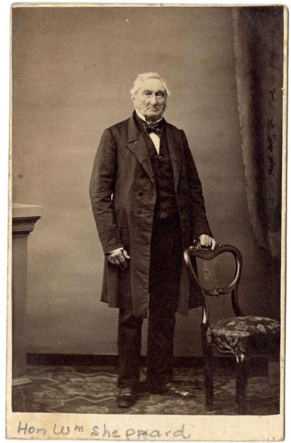 Black and white photograph of a man standing behind a chair, wearing a long black coat.