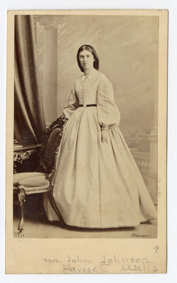 Black and white photograph of a woman standing behind the chair of a carved wooden desk, wearing a long dress with puffy sleeves and a net in her hair.