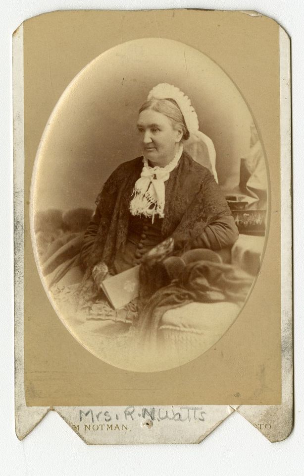 Photograph of a seated woman holding a book on her lap. She is wearing a lace shawl, a scarf around her neck, and a hat.