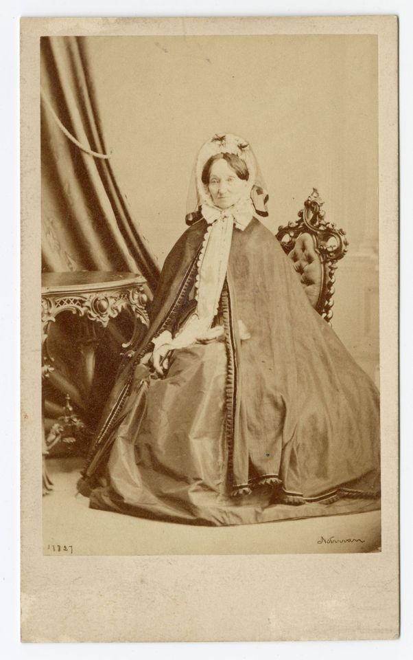 Black and white photograph of a woman sitting at a carved wooden desk, dressed in long garments decorated at the edges and wearing a hat.