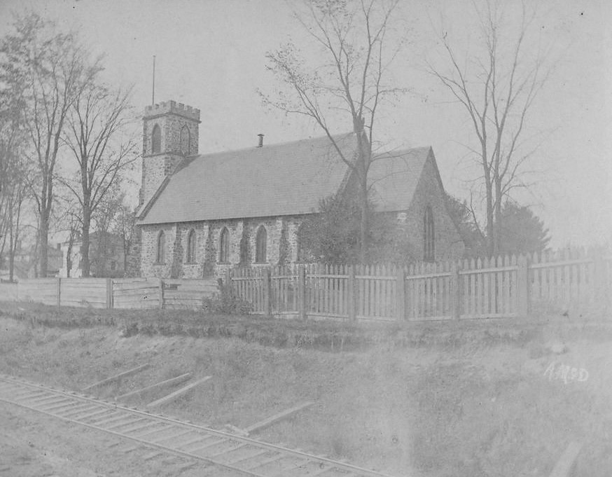Black and white photograph of St. George’s Anglican Church. It is a stone church with large stained-glass windows and an imposing steeple at the front. It is surrounded by a few trees and is located by the railroad tracks.