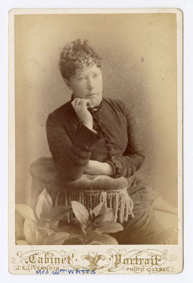 Black and white photograph of a seated woman posing, leaning on a cushion, wearing a long dress with sleeves decorated with buttons.