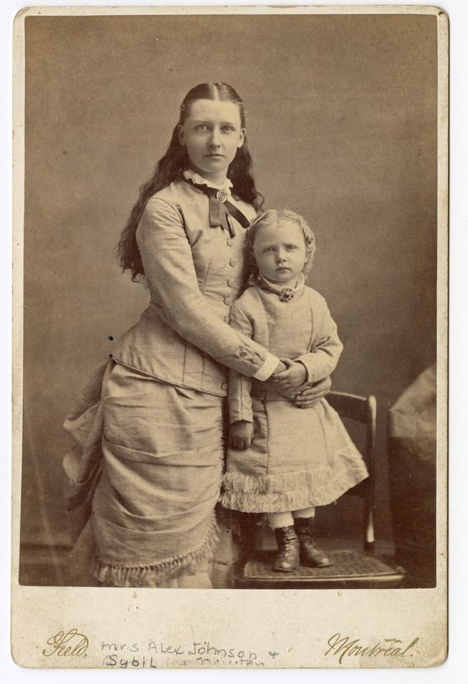Black and white photograph of a woman and a young girl, both standing, dressed in long ribbon-adorned dresses with a brooch at the neck. The child is standing on a chair to be at the same level as her mother.