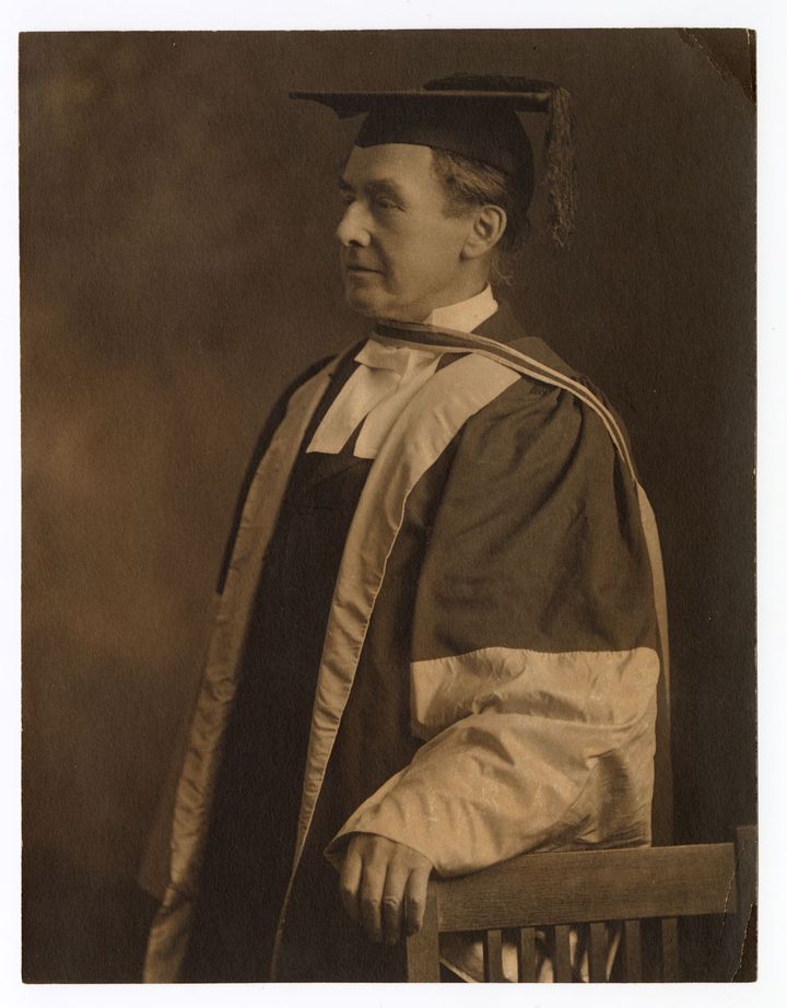 Black and white photograph of a man dressed in a robe and wearing a mortarboard, standing and leaning on a chair.