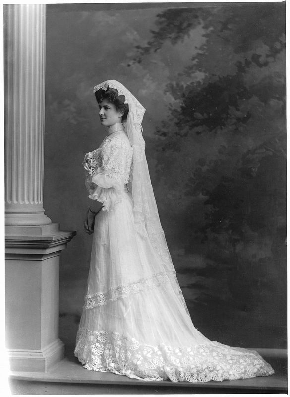 Black and white photograph of a woman posing near a column, wearing her wedding dress with lace and a veil.