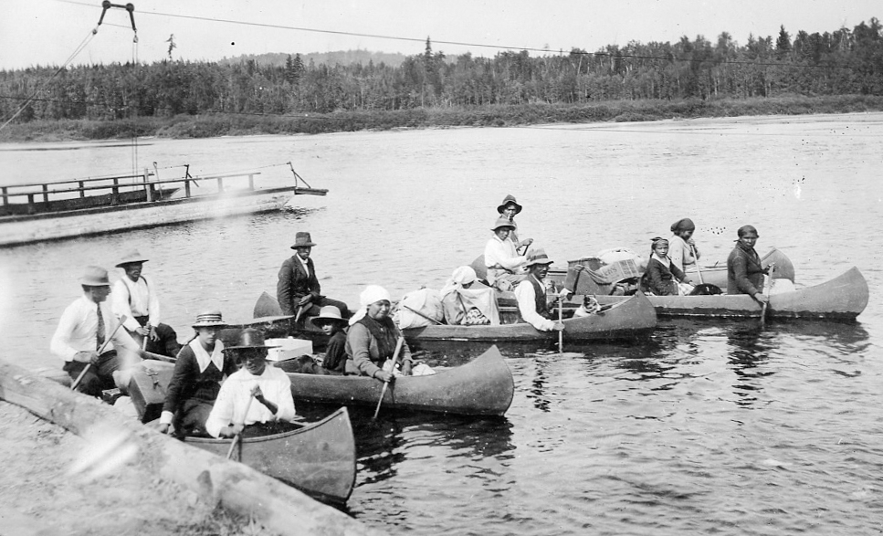 Indigenous people are sitting in five canoes filled with belongings on the river