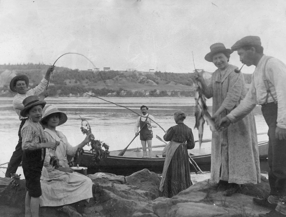 A family is fishing on the banks of the Saint-Maurice. A young boy is holding a fishing rod and a woman is holding up several fish.